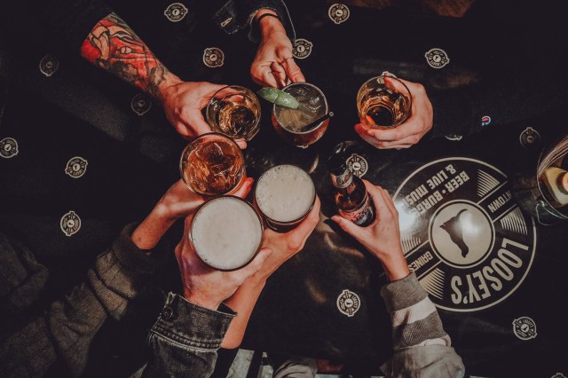 Image of Many Hands holding Cocktails and Pints of Beer over a table with the Loosey's Logo. For decorative Purpose only.